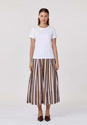 Femme debout, portant un t-shirt blanc uni à manches courtes, un pantalon large rayé marron et blanc, et des chaussures marron pointues.