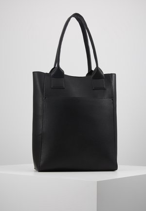Schwarze, strukturierte Leder-Tasche mit zwei langen Griffen, aufrecht stehend auf einem weißen Sockel vor einem einfarbigen hellgrauen Hintergrund.