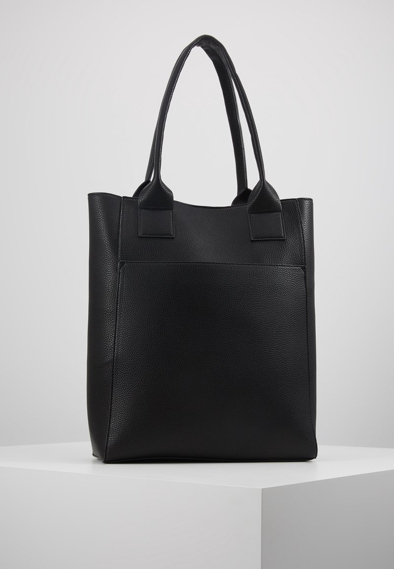 Sac cabas en cuir noir texturé avec deux longues anses, debout sur un piédestal blanc sur un fond gris clair uni.