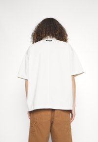 Bílá košile s krátkým rukávem, rovného střihu, s velkými rukávy a diskrétní etiketou na krku. Nosit se s hnědými cargo kalhotami.