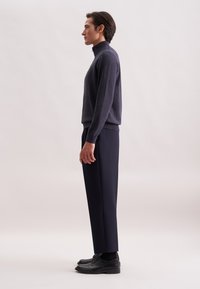 Uomo in maglione blu scuro, pantaloni neri su misura e scarpe eleganti nere in profilo contro uno sfondo chiaro e semplice.