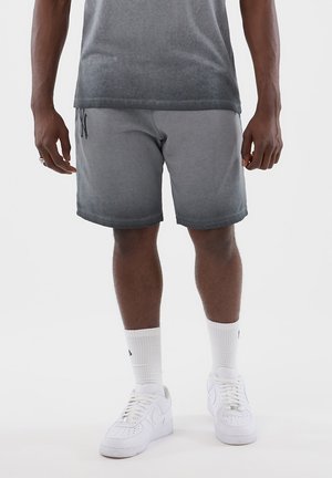 Hombre vistiendo pantalones cortos grises con efecto ombré, calcetas blancas hasta el tobillo y zapatillas blancas, posando contra un fondo claro y liso.