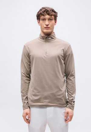 Ung mann iført en beige langermet topp med kvartglidelås og hvite bukser, stående mot en ensfarget lys bakgrunn.