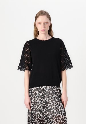 Haut en tricot noir avec des manches en dentelle, un décolleté festonné et un ourlet côtelé. Porté sur une jupe transparente à imprimé léopard, mettant en valeur un look superposé.