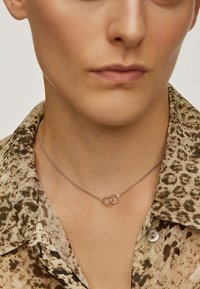 Collar de cadena de plata con dos anillos entrelazados; uno pulido en suave acabado, el otro con un diseño texturizado. Llevado sobre una camisa de estampado.
