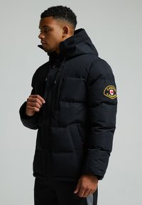 Schwarze Pufferjacke mit hohem Kragen, Reißverschluss vorne und mehreren Taschen. Mit einem Logo-Patch am linken Ärmel und einer matten Oberfläche.