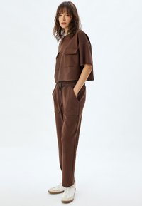 Bruine cropped t-shirt met twee zakken aan de voorkant, gecombineerd met bijpassende joggers met een elastische tailleband en zijzakken, gedragen met witte sneakers.
