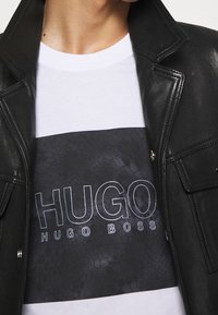 Černá kožená bunda přes bílý tričko s černým panelem s texturovaným vzorem a bílým a šedým textem "HUGO HUGO BOSS".