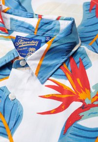 Camisa de manga corta con cuello en blanco, decorada con un vibrante estampado floral tropical en azul, rojo y naranja. Presenta un cierre de botones en el frente y una etiqueta con la marca.