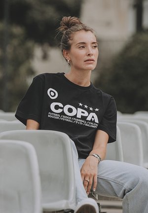 Zwarte T-shirt met korte mouwen met "COPA" in grote witte letters, met sterren en verschillende talen eronder. Gepaard met lichtblauwe spijkerbroek.