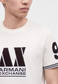 Νεαρός άνδρας με λευκό t-shirt Armani Exchange με έντονο μαύρο λογότυπο και αριθμό 9 στο μανίκι, σε απλό φόντο.