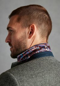 Blazer gris à motif chevrons avec un col bleu. Doublure bordeaux à motif floral émerge du col, mettant en valeur des détails complexes.