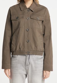 Veste en toile marron avec un col, deux poches poitrine et des poches latérales. Présente des fermetures à bouton et une taille élastique pour un ajustement optimal.