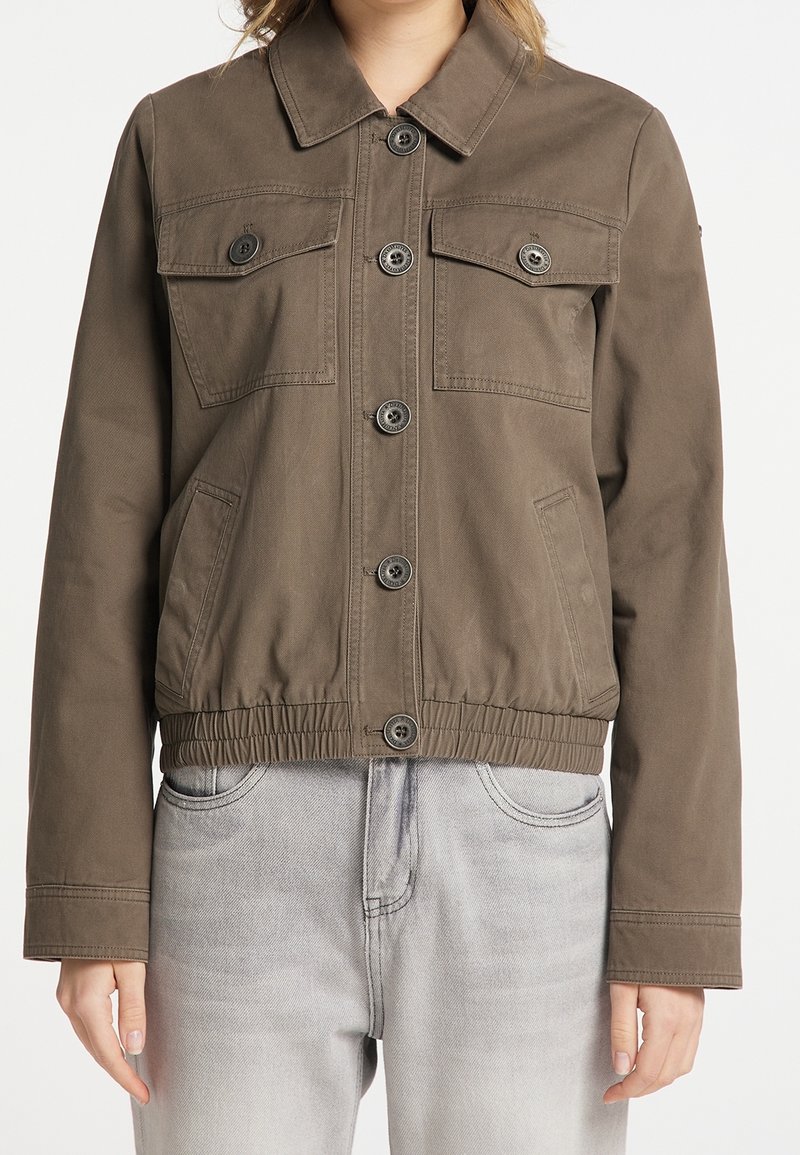 Veste en toile marron avec un col, deux poches poitrine et des poches latérales. Présente des fermetures à bouton et une taille élastique pour un ajustement optimal.