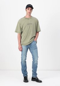 Olive zelené oversized tričko s nápisom "HUGO" v sivej farbe, modré džínsy a čierne topánky. Oblečenie je jednoduché a neformálne, s krátkymi rukávmi.