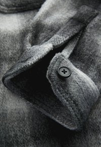Grijze wollen shirtkraag met een gestructureerd oppervlak, voorzien van een zwarte knoopdetail, en toont een verlooppatroon in donkerdere en lichtere tinten.