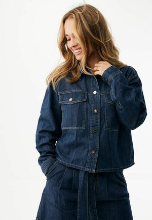 Usměvavá žena s blond vlasy, oblečená v tmavě modrém džínovém kabátku a odpovídající sukni, jedna ruka v kapse a druhá u límce.