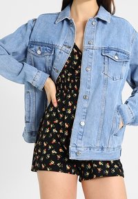 Chaqueta vaquera azul claro con un diseño clásico, cierre de botones, bolsillos en el pecho y un corte holgado, combinada con un mono negro de flores.