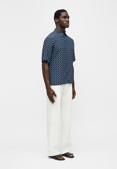 Moški stoji v modri srajci s kratkimi rokavi z vzorcem, belih širokih hlačah in temnih sandalih na enobarvnem svetlem ozadju.