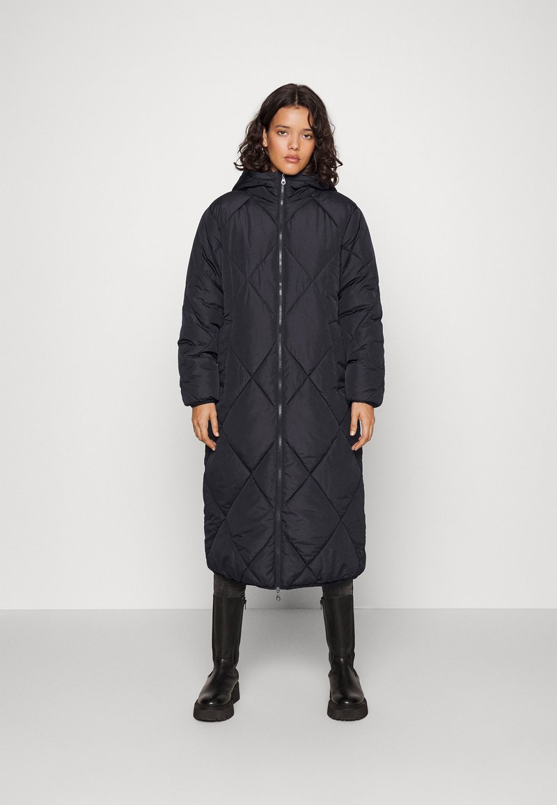Manteau matelassé noir, long, avec capuche, présentant une fermeture éclair à l'avant, un motif en diamant et des manches oversized. Porté avec des bottes noires montantes.