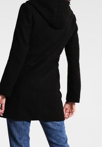 Abrigo negro con capucha y un diseño ajustado, de mangas largas y tejido texturizado. Presenta un corte a media longitud y detalles de costura vertical en la parte trasera.