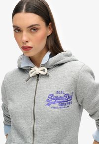 Felpa grigia con zip frontale, cordino bianco e logo ricamato viola. Presenta un colletto a strati di un blu chiaro. Tessuto morbido e texture.