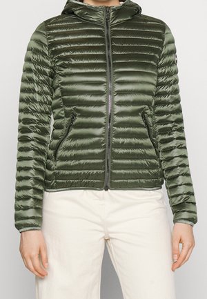 Femme portant une doudoune ajustée brillante vert olive avec capuche et poches zippées, associée à un pantalon blanc cassé.