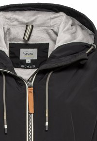 Schwarze Jacke mit einer fleecegefütterten Kapuze, ausgestattet mit einem silbernen Reißverschluss, beigen Kordeln und einem Etikett mit der Aufschrift "recycelt."