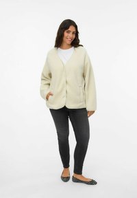 Cárdigan de forro crema con escote en V, manga larga y bolsillos frontales, combinado con jeans oscuros y planos negros con estampado.