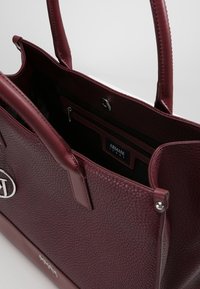 Bordowa torebka typu tote ze skóry z teksturowanym wykończeniem, podwójnymi uchwytami oraz zamykaną na zamek kieszenią wewnętrzną. Wnętrze z delikatnym oznakowaniem marki.