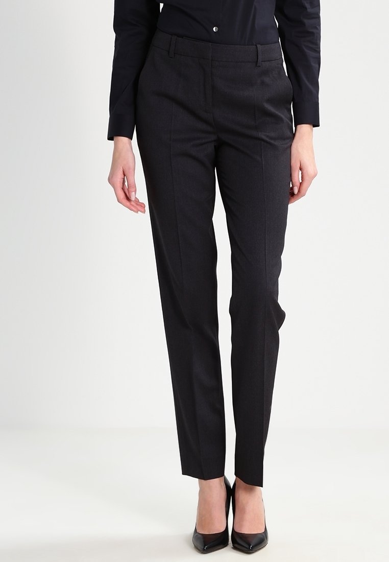 Femme portant un pantalon noir cintré et des talons hauts noirs pointus, debout devant un fond clair uni.