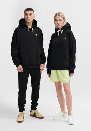 Zwarte hoodie met beige trekkoorden, gecombineerd met zwarte joggers en een lichtgroene miniboard rok. Sneakers in beige en wit.