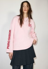 Hosszú ujjú világos rózsaszín ing piros "PURO SABOR" felirattal és logóval az elején. Sötét, pliszírozott szoknyával viselve. Simogató pamutanyag.