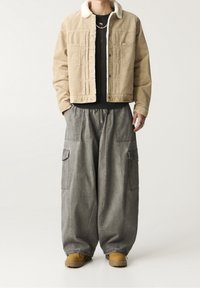 Tan kordflöjlskavaj med fårskinnskrage, bärs över en svart tröja, kombinerad med lösa grå cargobyxor och bruna stövlar.