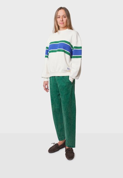 Witte sweatshirt met blauwe en groene strepen, gecombineerd met groene corduroybroek. De outfit wordt compleet gemaakt met donkerbruine instap schoenen.