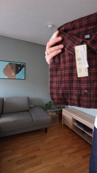 Raudonos ir juodos spalvos languota medvilninė marškiniai su sagomis ant apykaklės, ant kurios pritvirtinta etikėtė, laikoma priešais pilką sofą ir sienos dekoraciją.