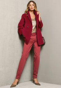 Bordeaux wollen jas met een brede kraag, gecombineerd met lichtroze broek met riem en bruine puntige hakken. Textuur gouden top eronder.