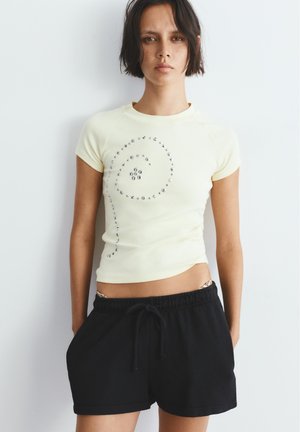 Femme portant un haut moulant à manches courtes jaune pâle avec un motif de clous en spirale et un short noir à cordons, debout devant un fond blanc.