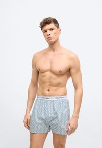 Άνδρας μοντέλο με σγουρά μαλλιά, φορώντα ς ανοιχτό μπλε κοντό εσώρουχο Calvin Klein με σχέδια, στέκεται μπροστά σε απλό λευκό φόντο.