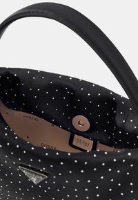 Schwarze Handtasche mit glatter Textur, innen mit beigem Stoff ausgekleidet und weißen Punkten verziert, ausgestattet mit einem Metall-Druckknopfverschluss und Markenlogo.