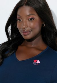 Laivastonsininen v-aukkoinen toppi pehmeästä kankaasta, jossa on vaaleanpunainen ja punainen sydänkuvio sekä teksti "Kiss me" vasemmassa rintapäässä.