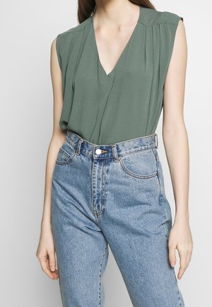 Kobieta w zielonej bluzce bez rękawów z dekoltem w serek, wpuszczonej do wysokich, jasnoniebieskich jeansów, stojąca z luźno opuszczonymi ramionami.