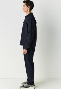 Chaqueta de mezcla de algodón azul marino con cierre de botones, un bolsillo en el pecho y pantalones a juego. Zapatillas grises y negras con cordones.