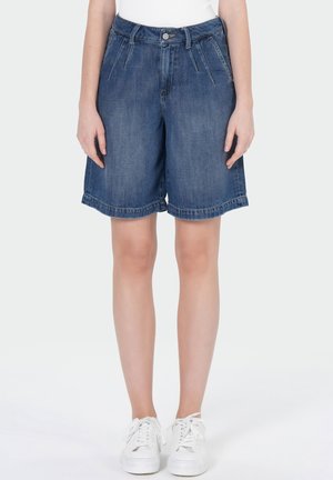 Personne portant un short en jean bleu longueur genou et des baskets blanches, debout avec les bras détendus le long du corps.