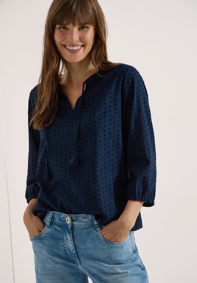 Jeune femme souriante, mains dans les poches, portant un chemisier bleu marine texturé avec des liens à pompons et un jean bleu clair sur fond uni.