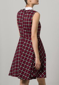 Šaty bez rukávů s červeným kostkovaným vzorem, bílým límečkem a přiléhavým trupem, který přechází do rozšířené sukně. Detail na zadním zipu.