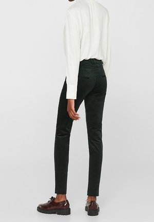 Personne portant un pantalon noir slim, une blouse blanche à manches longues et des chaussures marron à lacets, debout dos à la caméra.