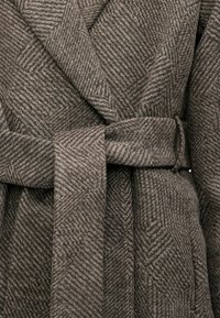 Close-up of brown herringbone wool coat with wide lapels and tied belt, showing detailed fabric texture and pattern.