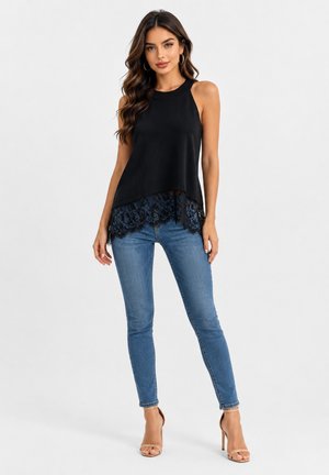 Vrouw draagt een zwart mouwloos topje met kanten zoom, blauwe skinny jeans en beige sandalen met hoge hak, staand tegen een simpele achtergrond.