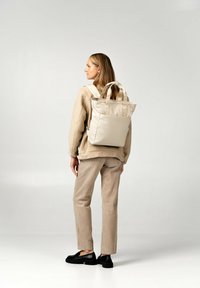 Beige tote bag som bärs som en ryggsäck, tillverkad av ett slätt material. Har två topphandtag och justerbara remmar. Enkel, strukturerad design.